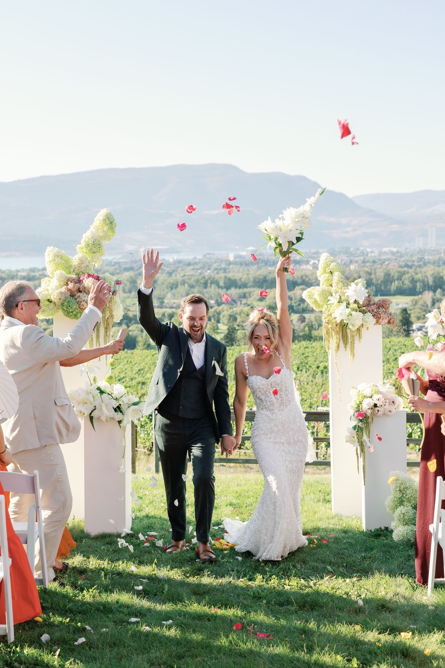 A joyful outdoor wedding celebration with guests throwing flower petals at a scenic mountain overlook on a sunny day.