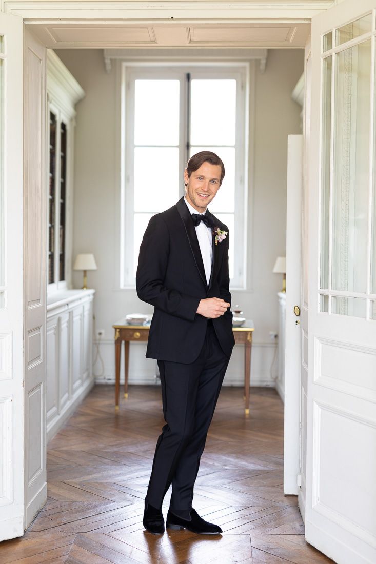 Full-length portrait of a groom in a classic tuxedo standing in a bright white hallway.