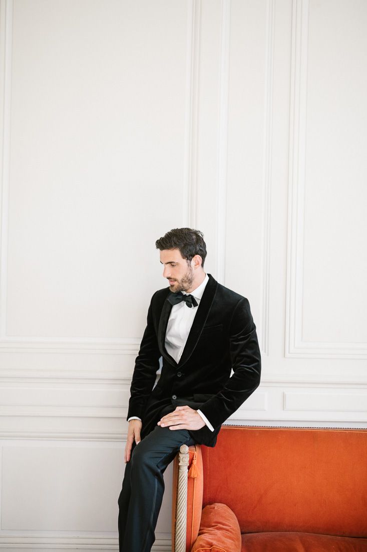 Groom in velvet tuxedo sits casually on orange couch against white wall.
