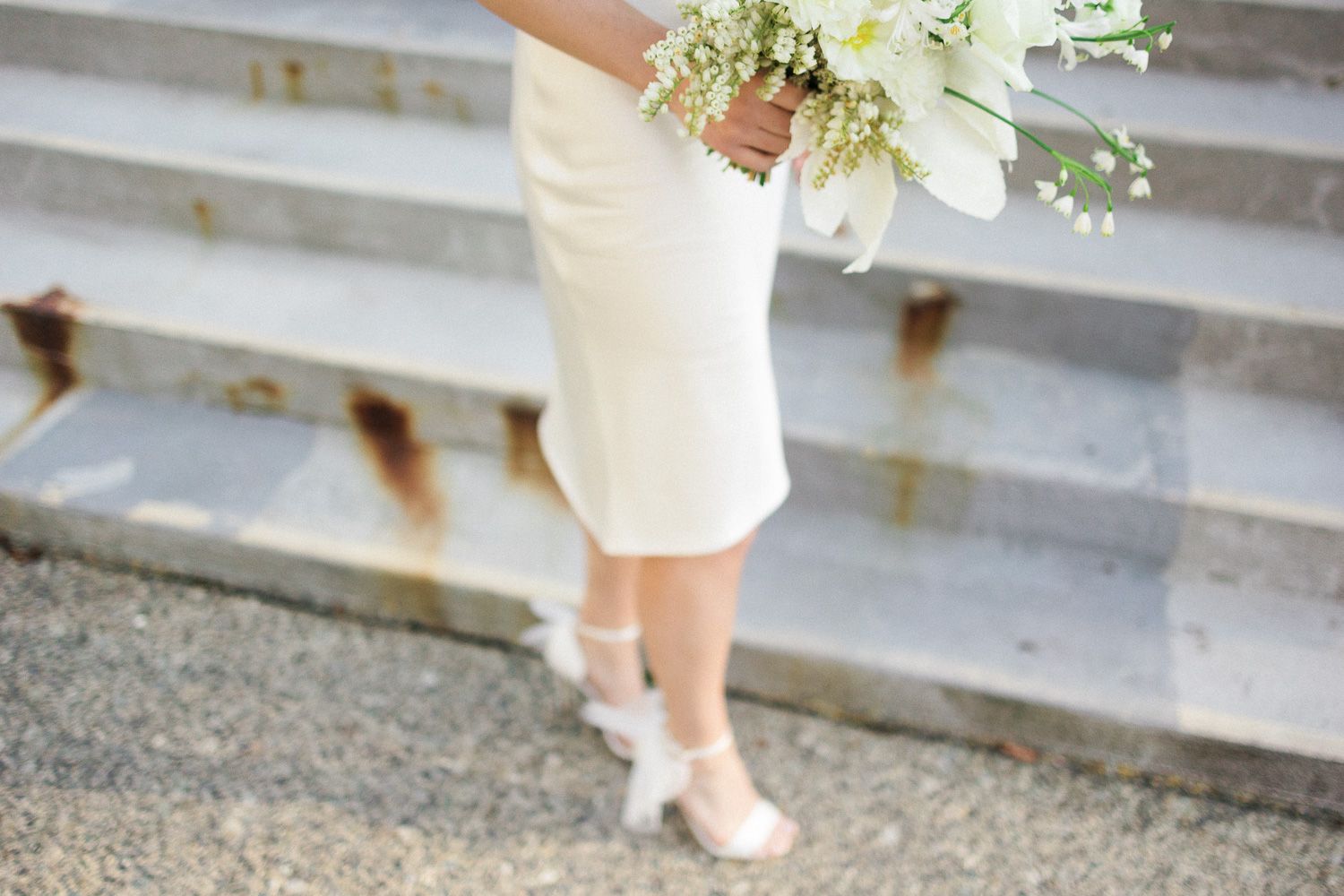 Elegant white wedding dress and bouquet detail on stone steps with delicate floral arrangement.