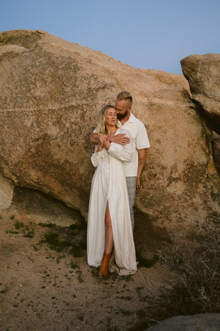 A couple poses against a large desert boulder during golden hour.