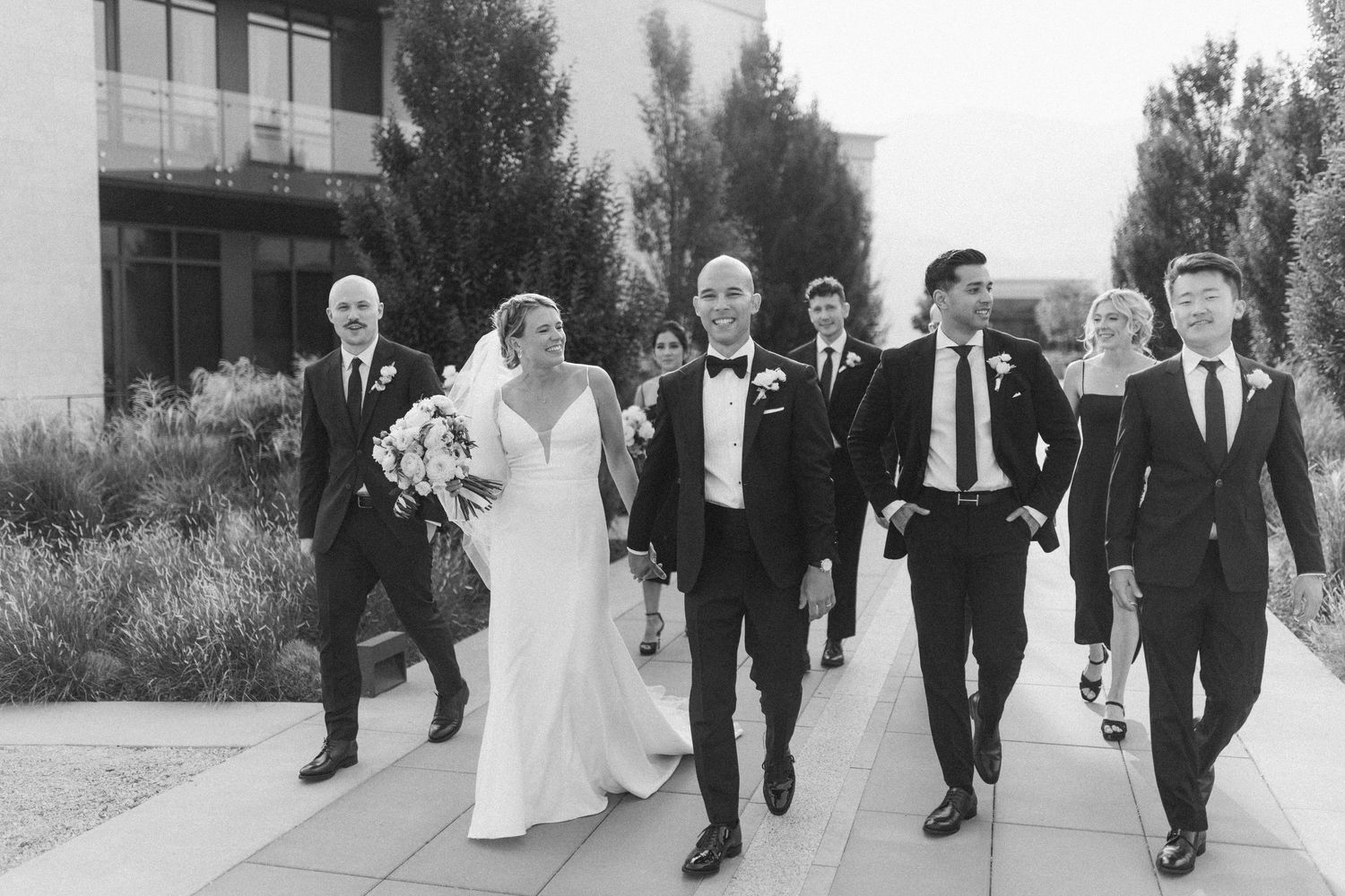 Wedding party walking and laughing together along an outdoor pathway in black and white.