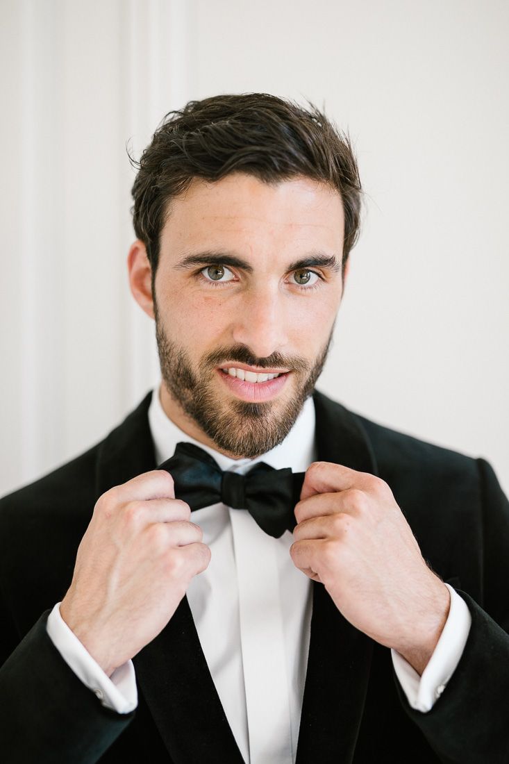 Groom in black tuxedo adjusts bow tie while smiling.