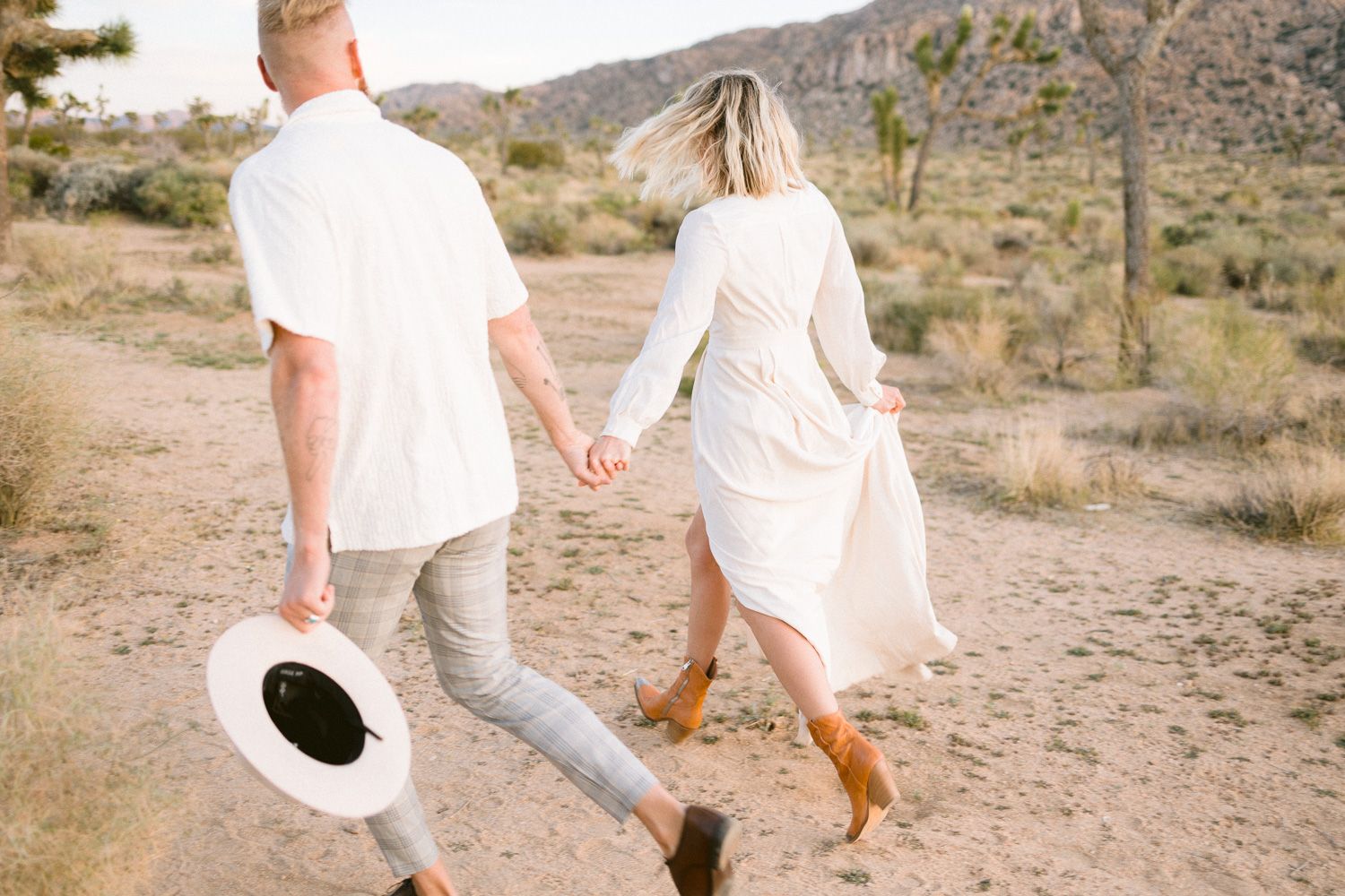 Couple walking hand in hand through a desert landscape with Joshua trees at sunset.