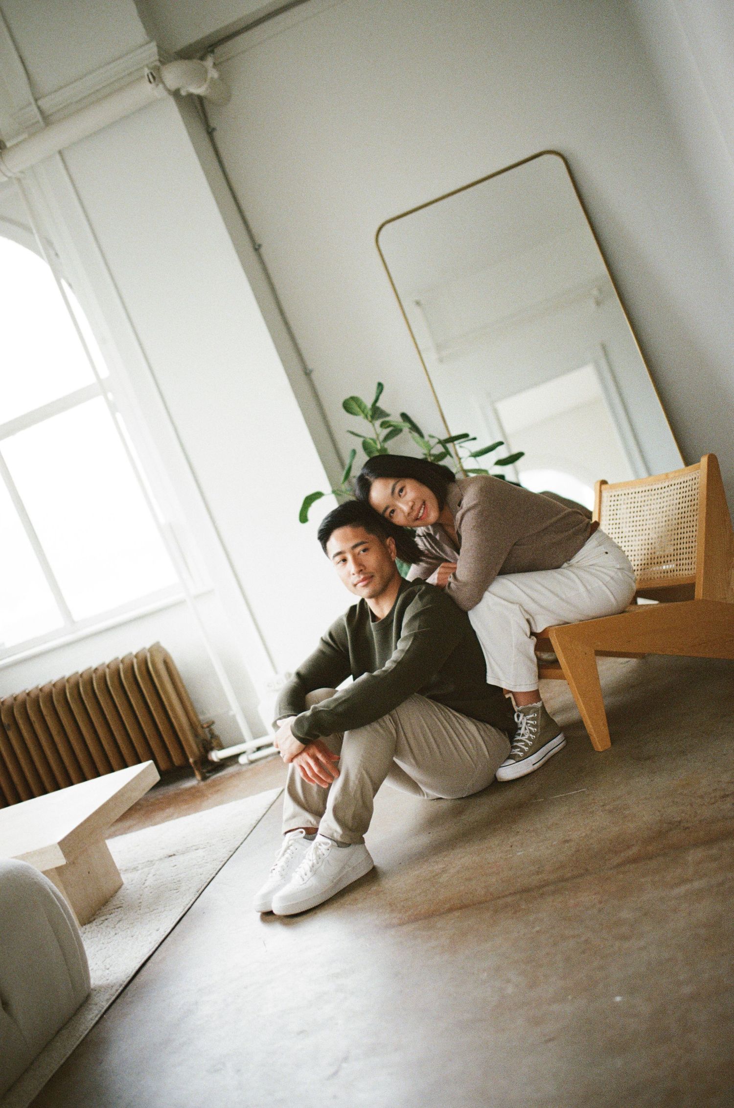 Two people in casual attire relaxing together in a minimalist room with a mirror and houseplant.
