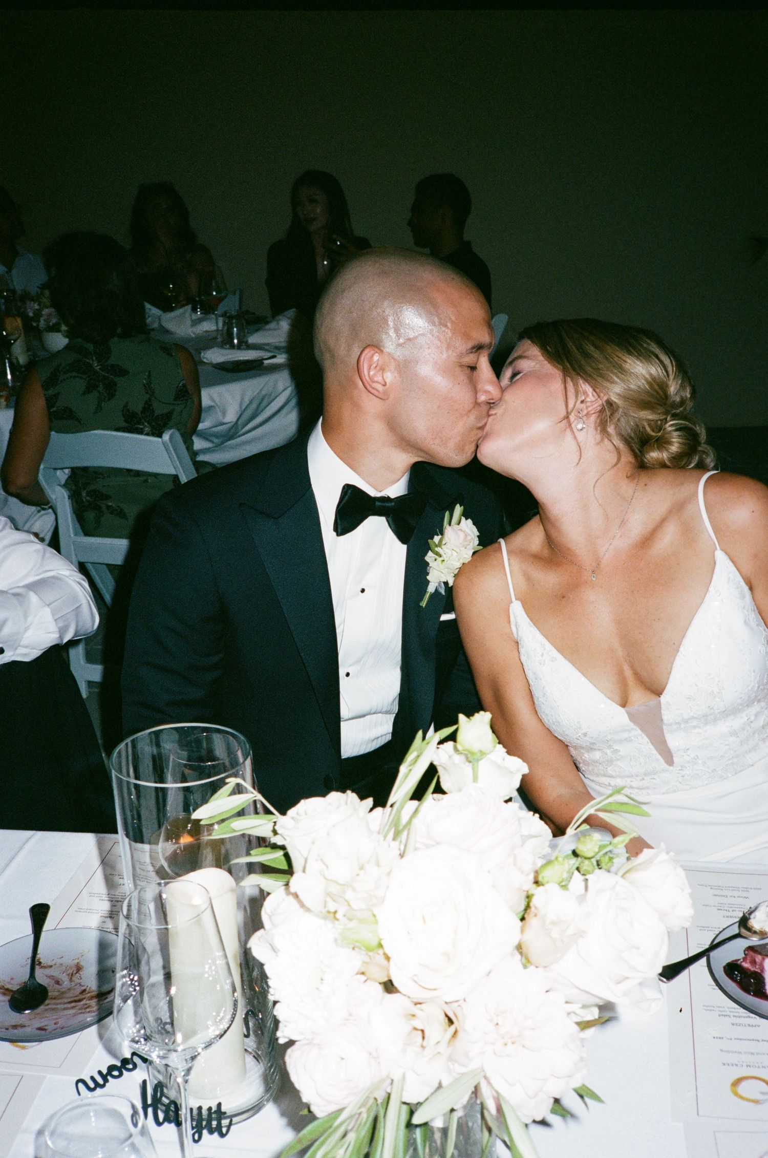 A couple shares a romantic kiss at their wedding reception table decorated with white floral centerpieces.