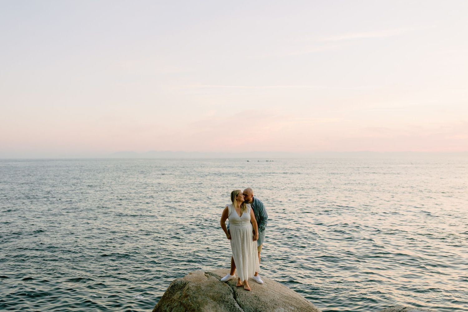 A couple in wedding attire stands together on a rocky outcrop overlooking the ocean at sunset.