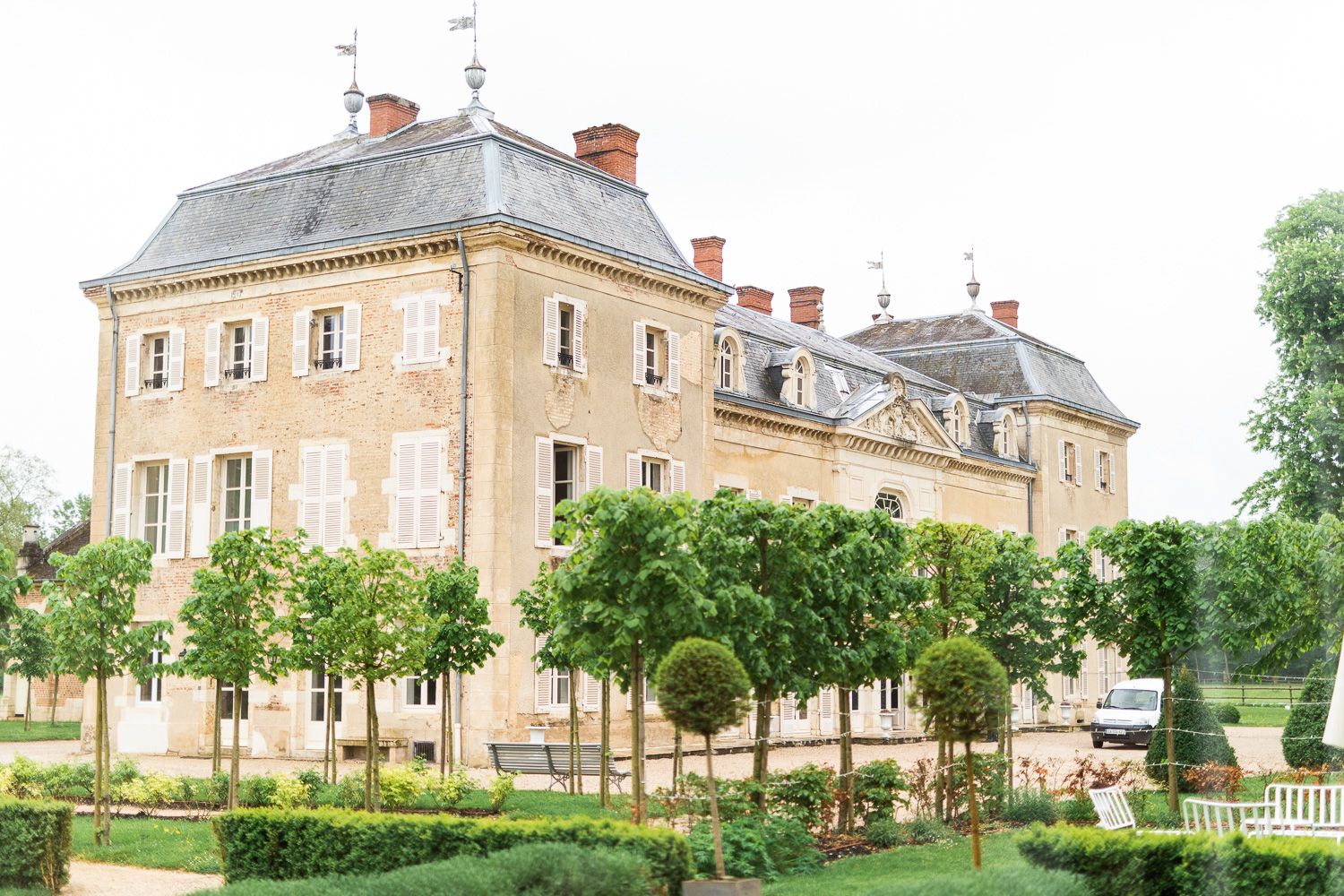 Stately beige stone chateau with mansard roof surrounded by formal gardens and trees.