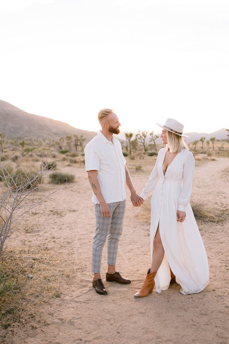 A couple in contrasting white outfits stands together on a desert path surrounded by cacti.