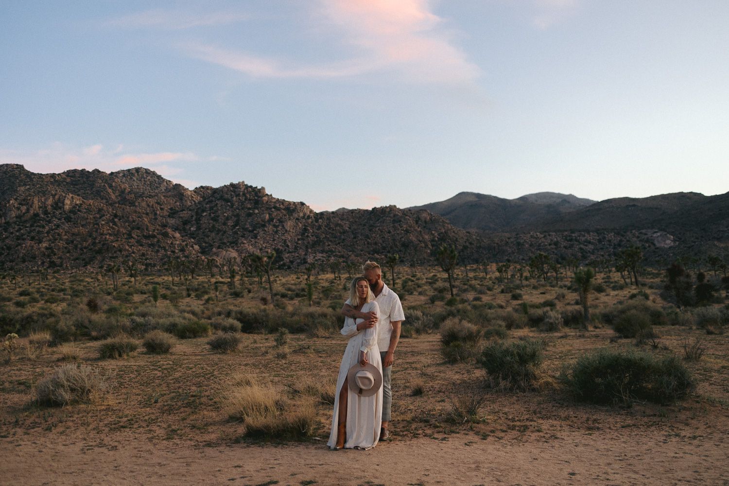 Two people share an intimate moment in the desert with mountains in the background.