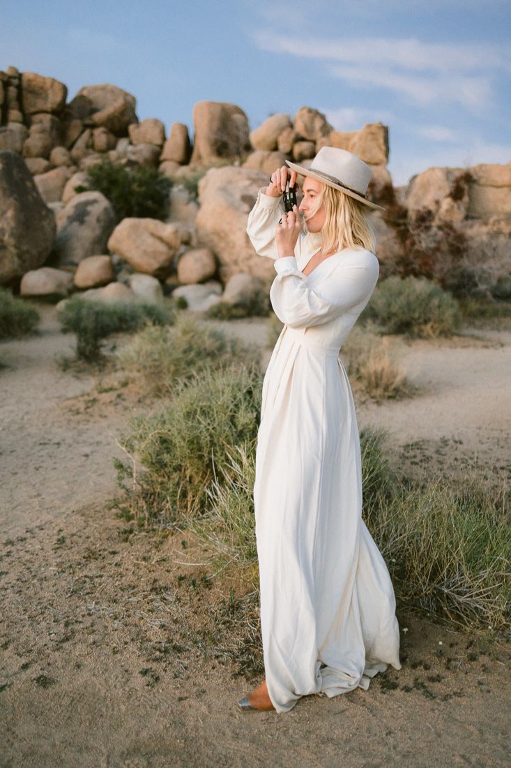 Woman in white maxi dress and hat poses against boulder formations in desert landscape.