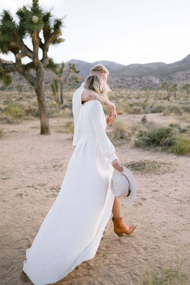 A bride in a flowing white dress holds a hat while walking through a Joshua tree desert landscape.