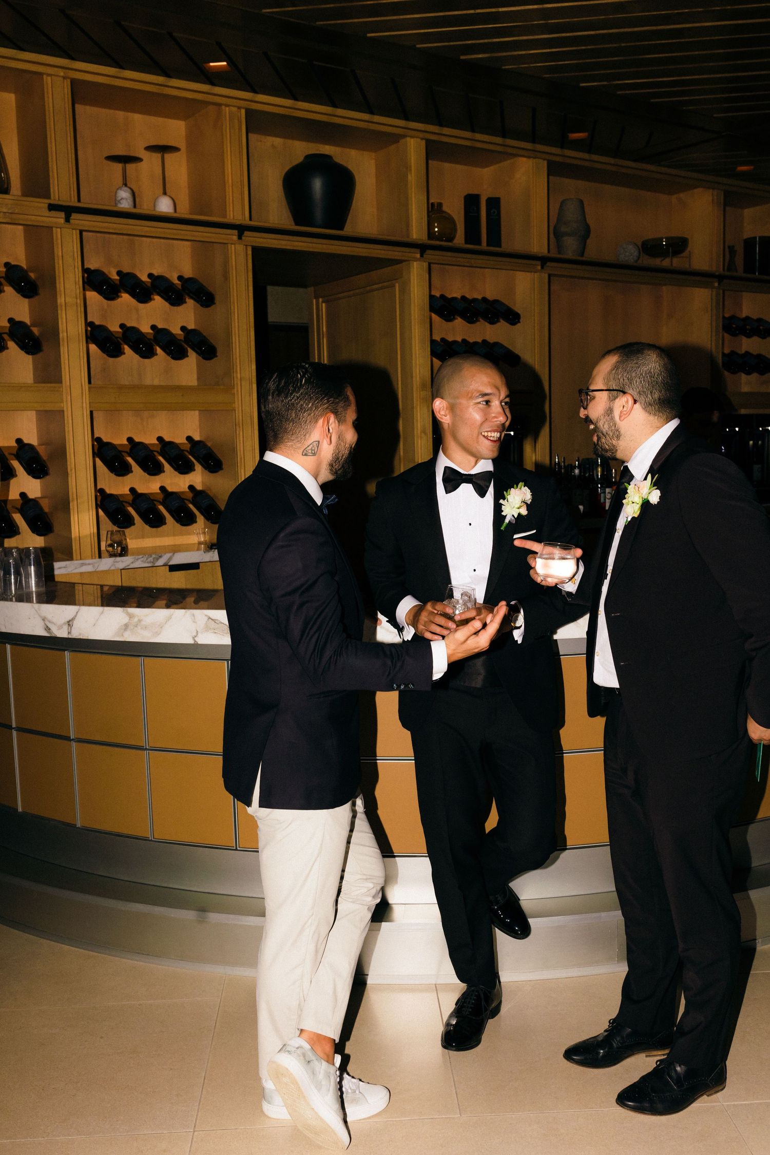 Formal-dressed guests in tuxedos conversing at a wine cellar reception with wooden wine racks in background.