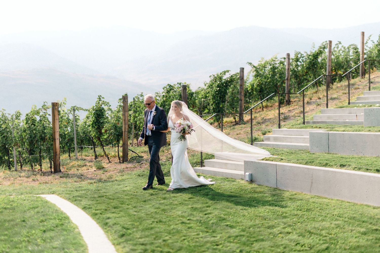 Wedding couple walking along a scenic hillside vineyard path surrounded by greenery.