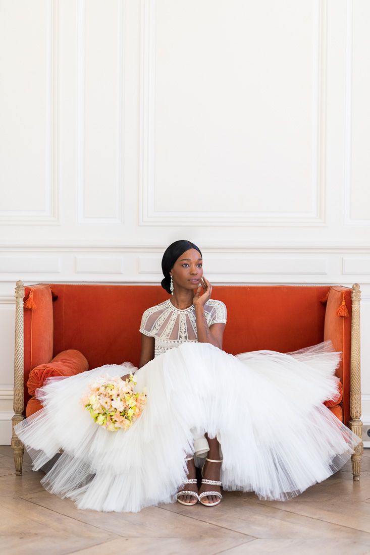 Bride wearing white lace dress relaxes on vintage orange sofa with floral bouquet.