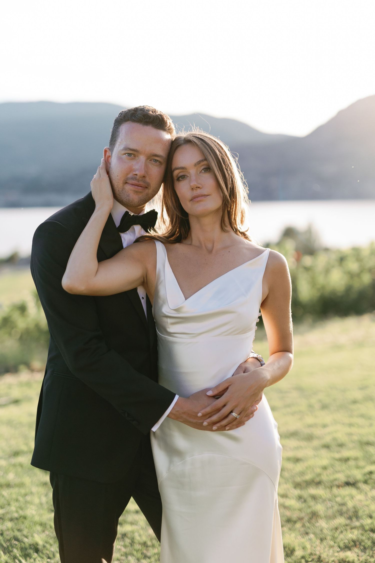 Bride and groom embrace outdoors at golden hour, she in white satin gown, he in black tuxedo with bow tie.