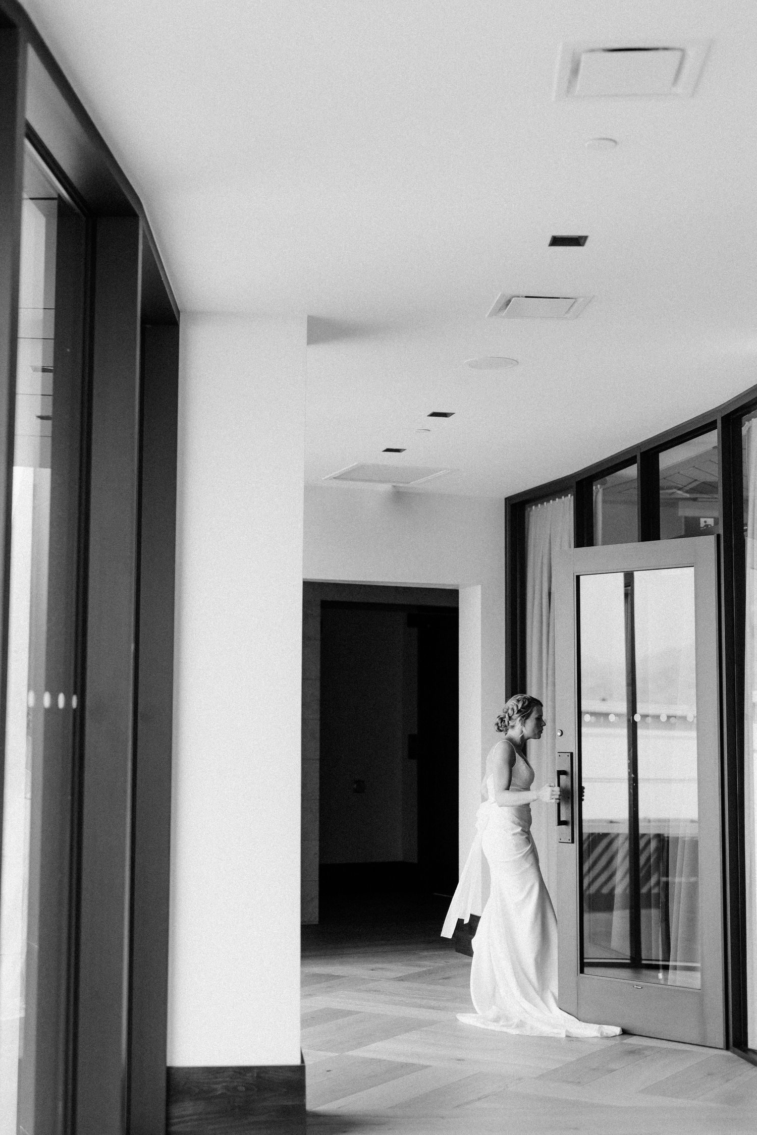 An elegant figure in a white gown stands thoughtfully in a modern hallway.