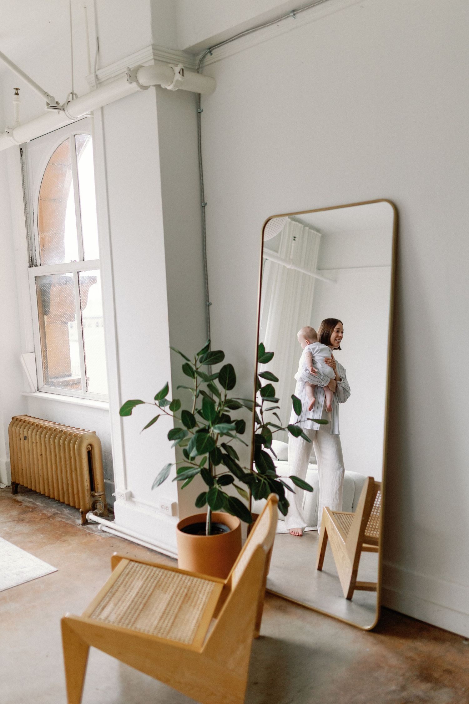 A minimalist white room with wooden chair, leaning mirror, and potted plant by sunlit window.