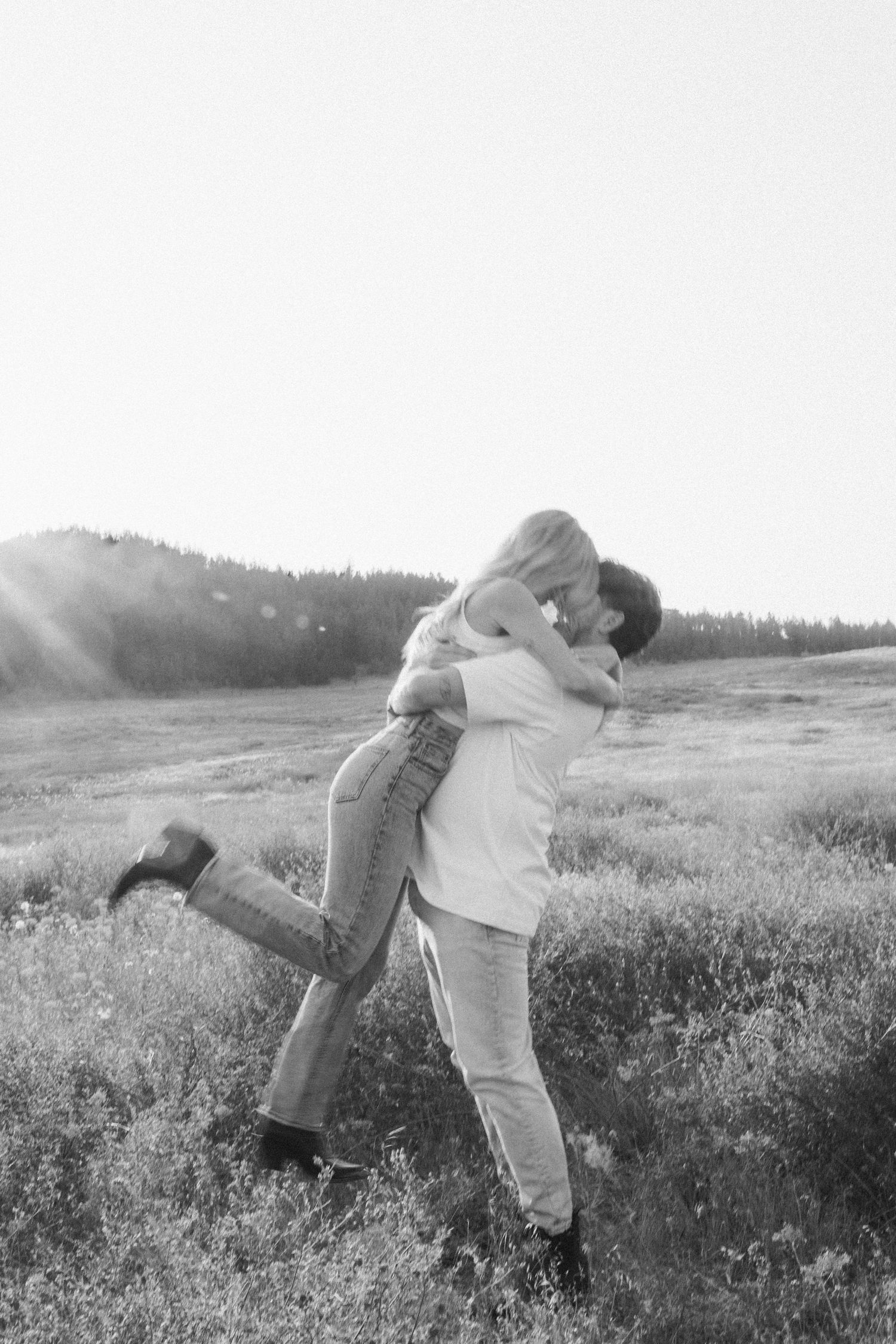 Couple embracing in a sunlit meadow with mountains in the background captured in black and white.