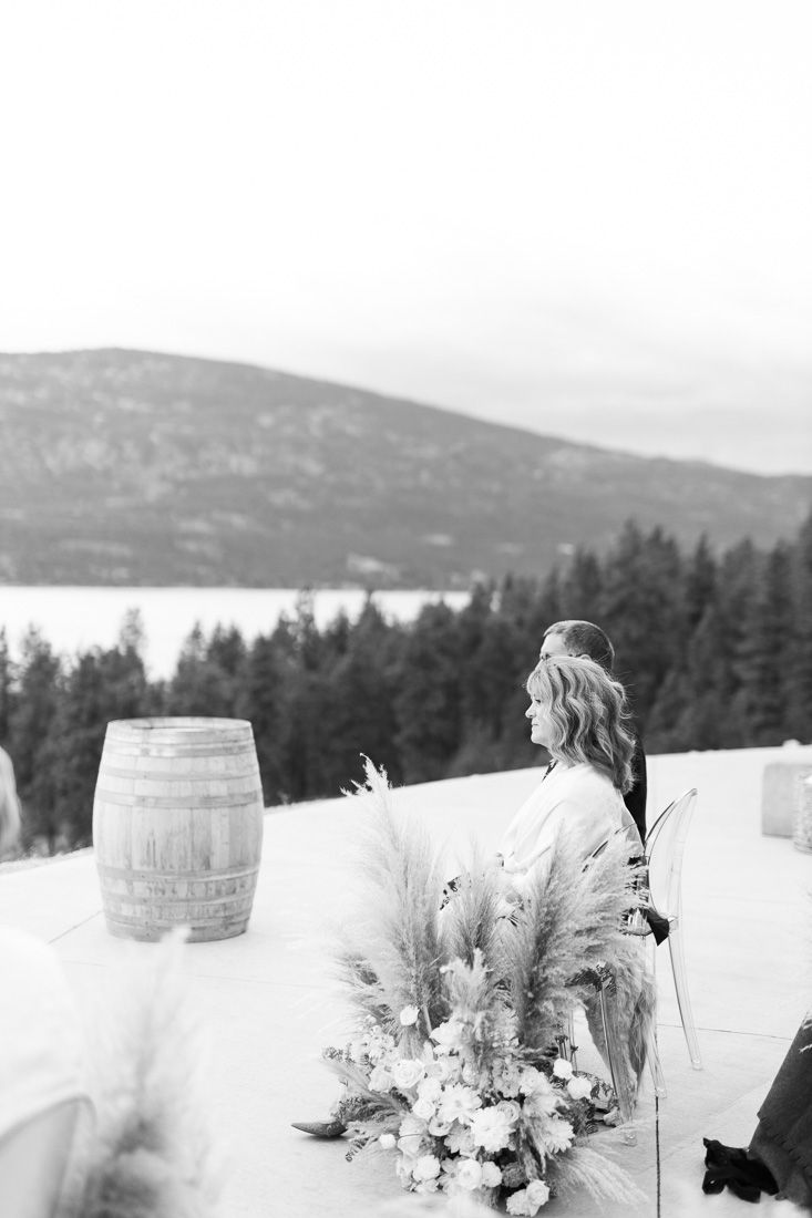 A rustic mountain wedding setting with wine barrel and floral arrangements against a lake backdrop.