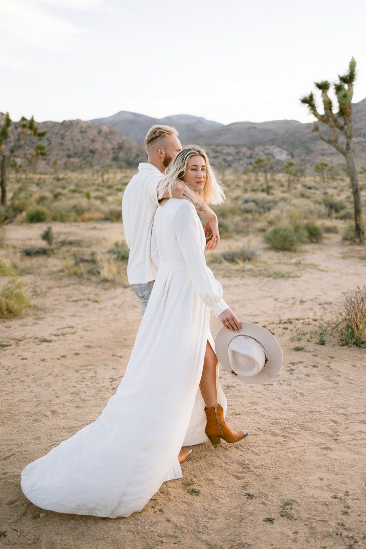 A romantic desert photoshoot featuring flowing white dress and brown boots against a backdrop of Joshua trees and mountains.