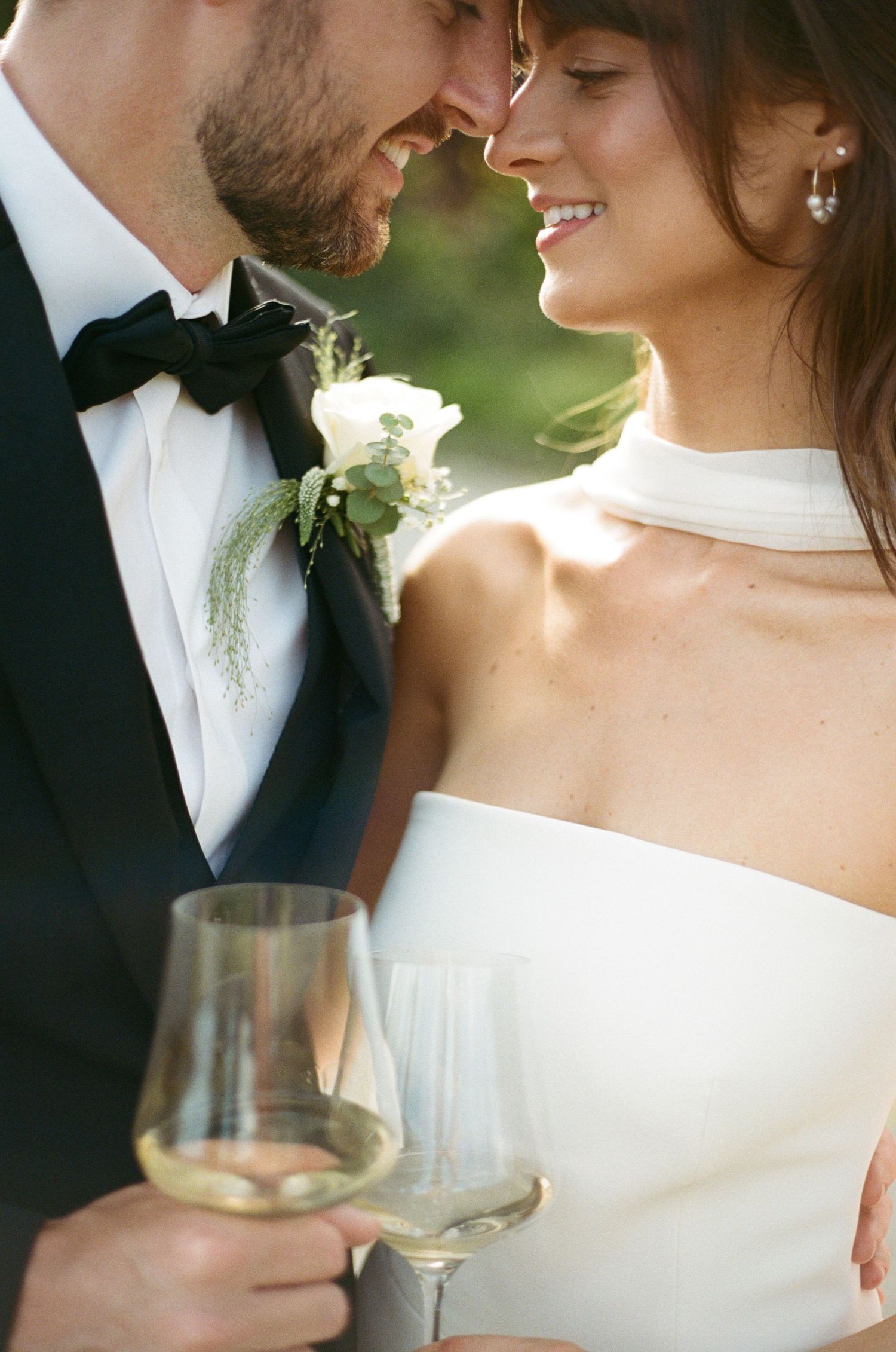 Romantic wedding couple shares intimate moment while holding wine glasses in elegant outdoor setting.