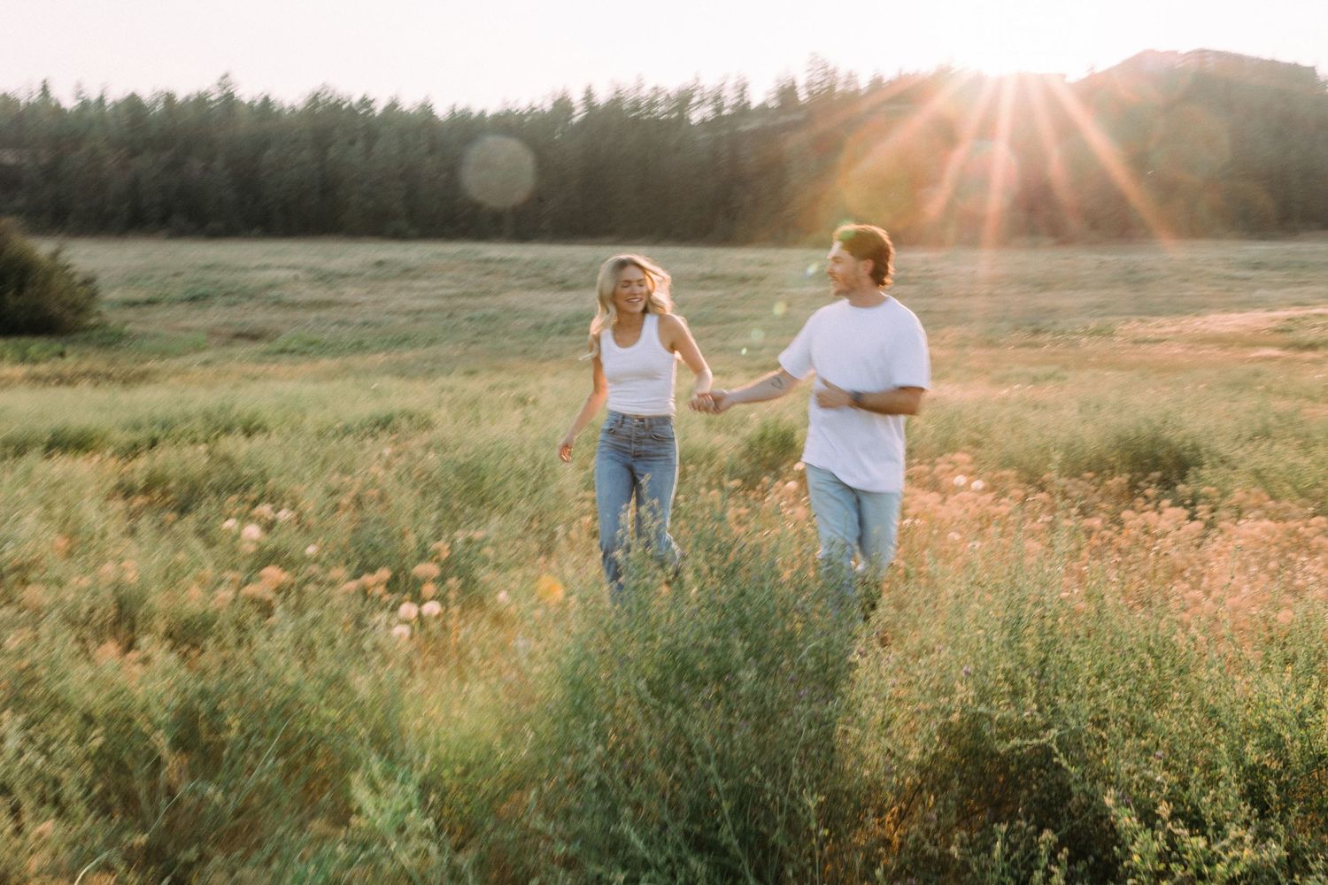 Couple walking through meadow at sunset with sun flare and mountain backdrop.