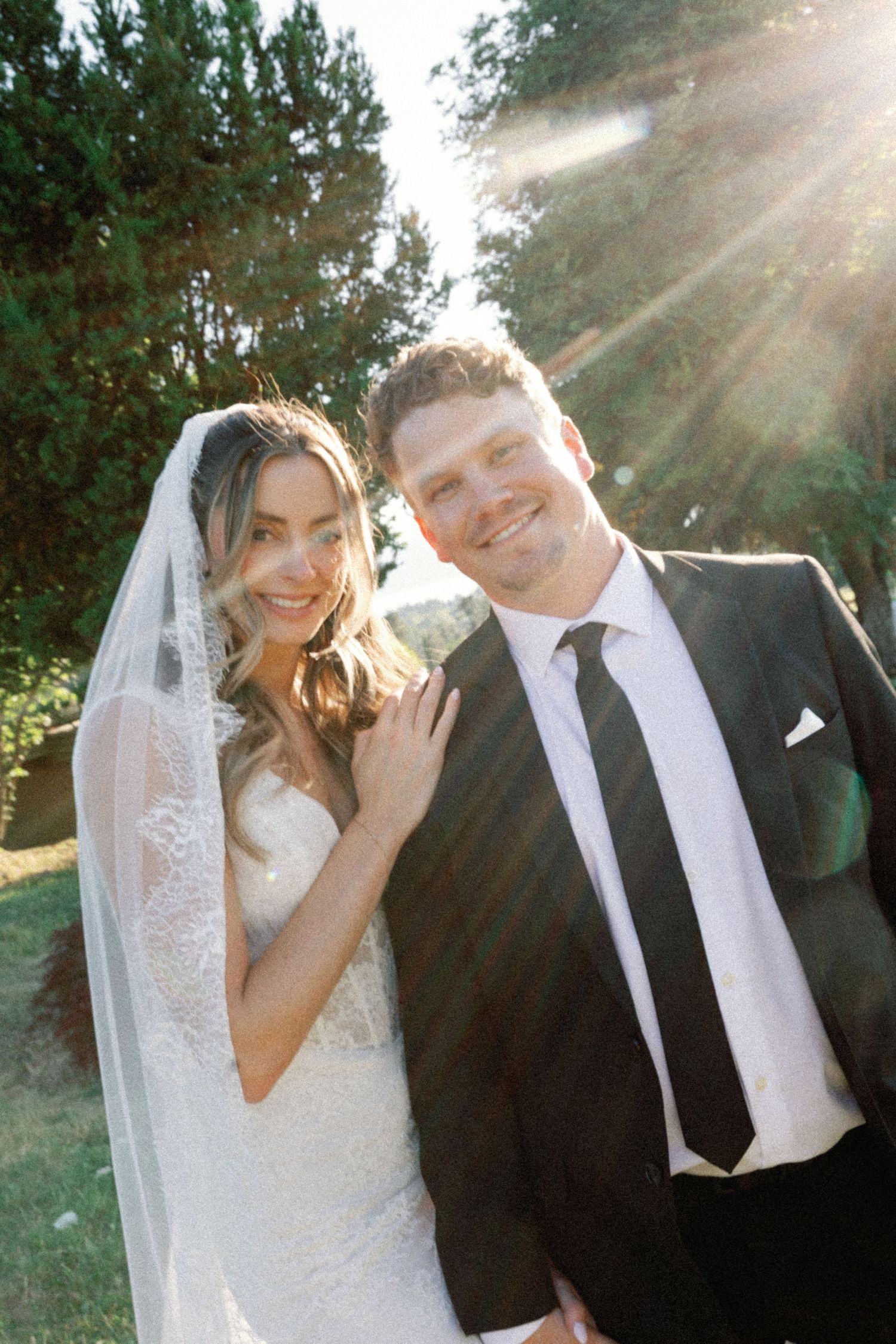 A couple poses together outdoors on their wedding day with sunlight streaming through trees in the background.