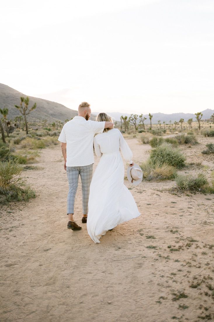 A couple walks hand in hand through a desert landscape dotted with Joshua trees at sunset.
