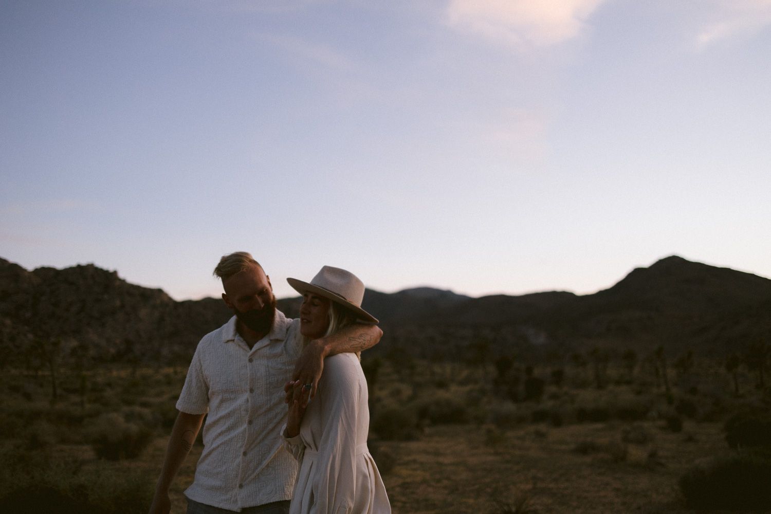 A romantic couple stands together in a desert landscape at sunset with mountains in the background.