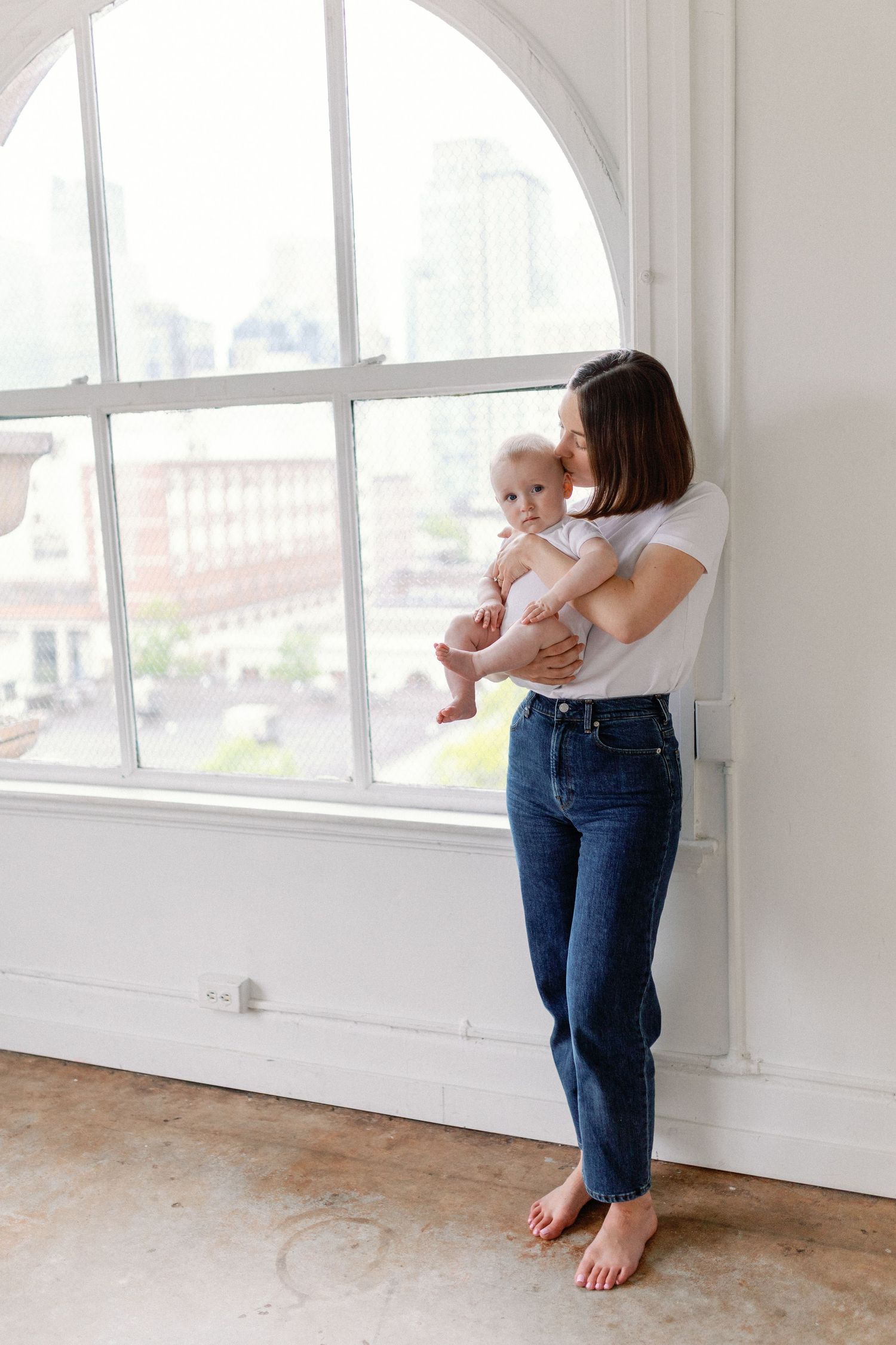 Parent plays with baby in bright sunlit room with large windows and bare wooden floors.
