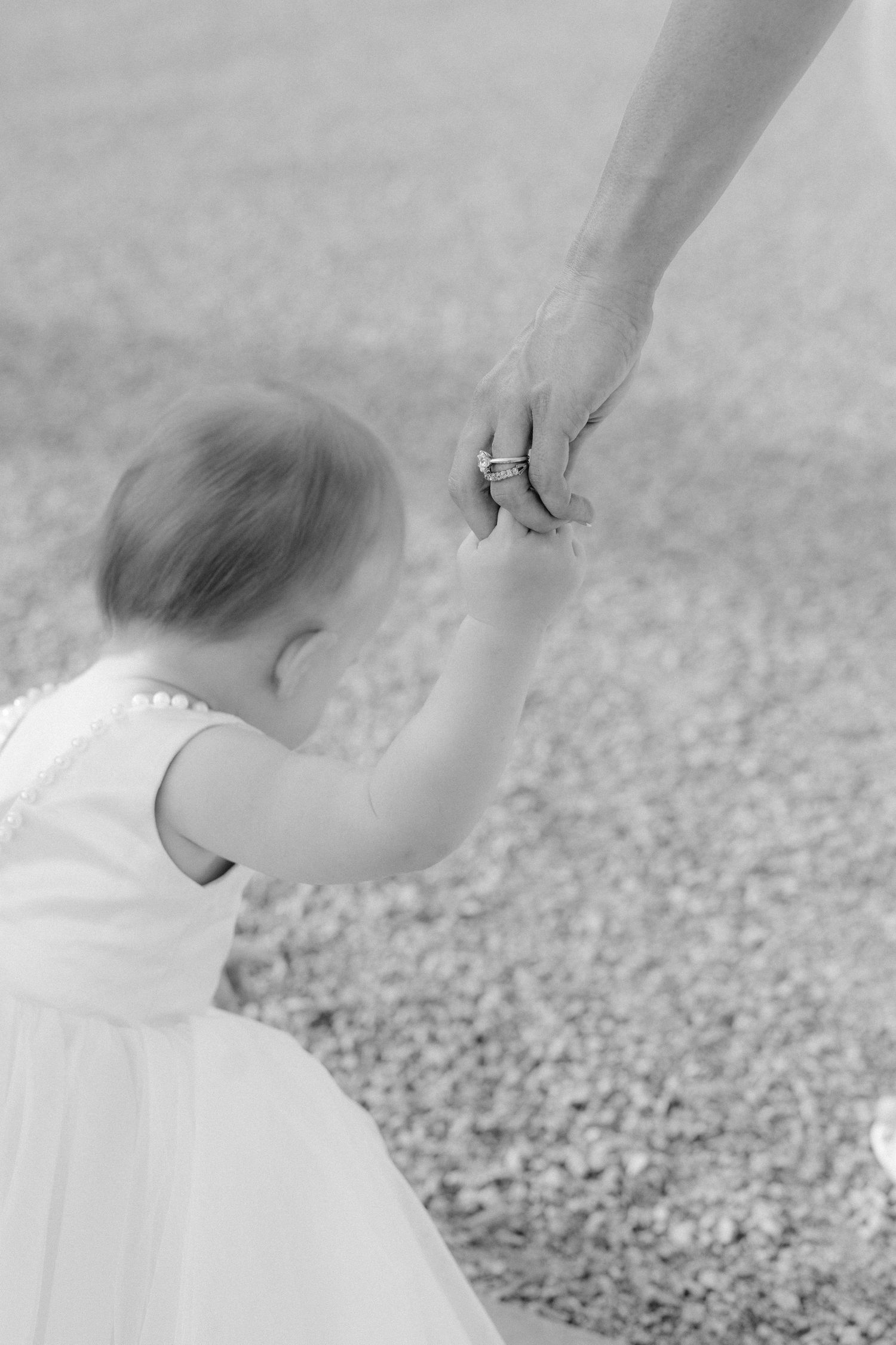 Black and white close-up of a small child in white dress holding an adult's hand with wedding rings.