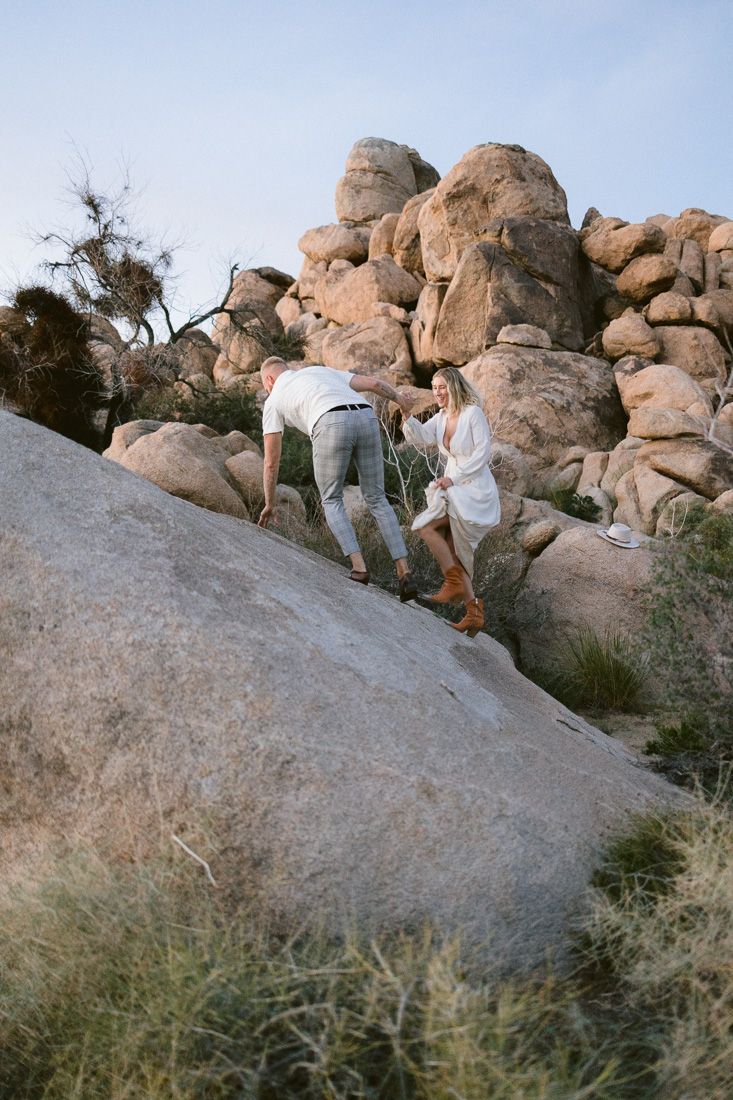 Couple climbing rocky terrain together in desert landscape at sunset.