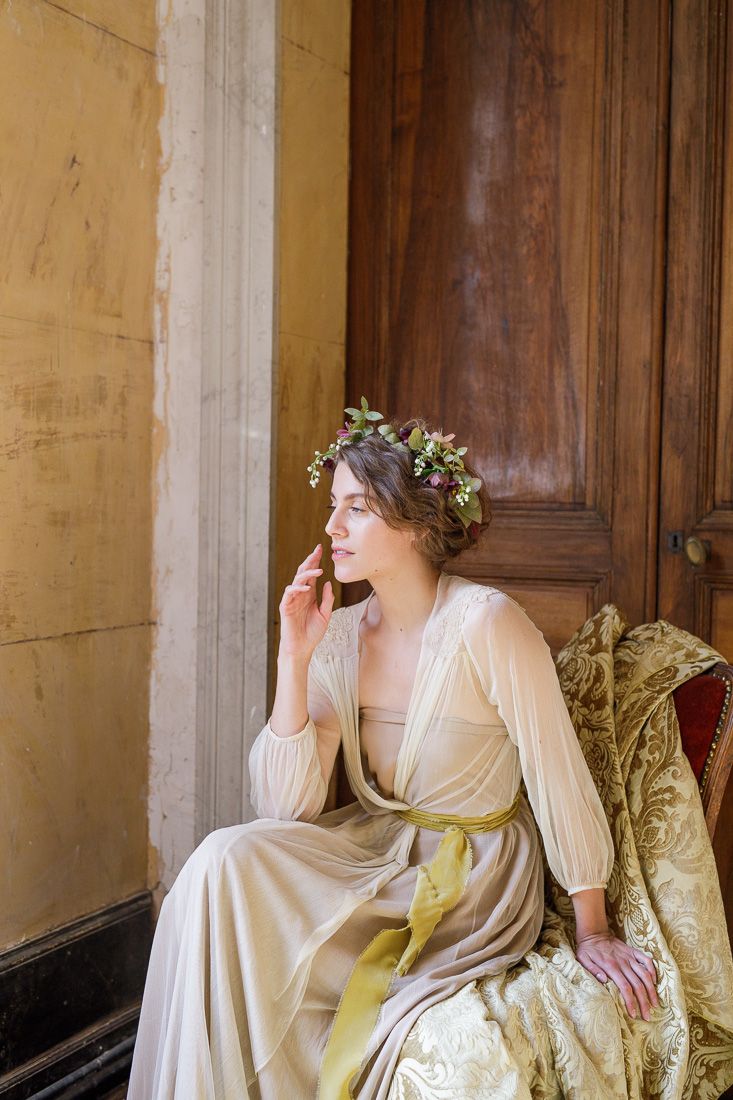 Ethereal portrait of someone in a vintage-style dress and floral crown seated by wooden doors.