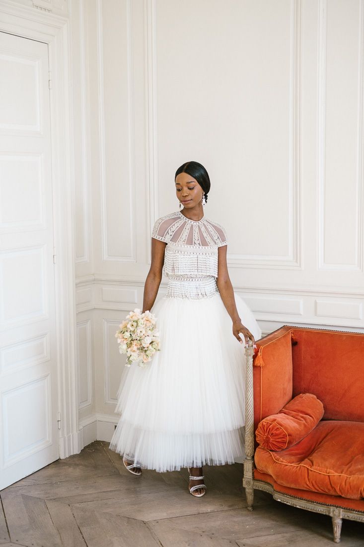 Bride wearing white tea-length gown with lace bodice poses next to orange antique sofa.