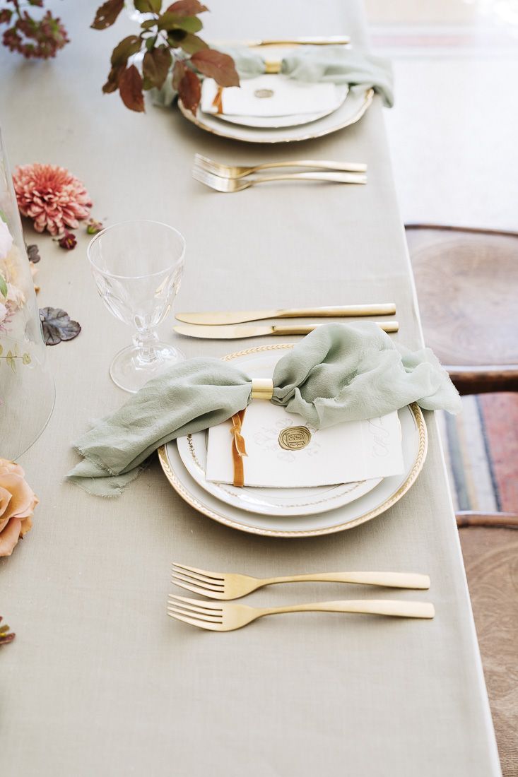 Minimalist place setting with sage napkin, gold flatware, and white plates on gray linen tablecloth.
