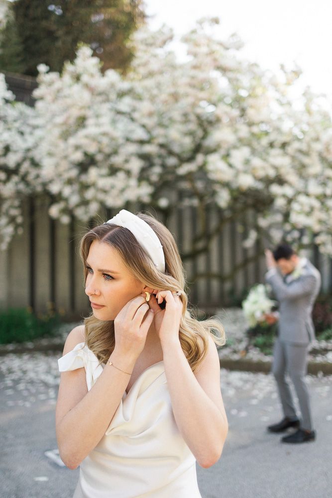 Bride adjusts earring in foreground with groom visible in background under flowering trees.