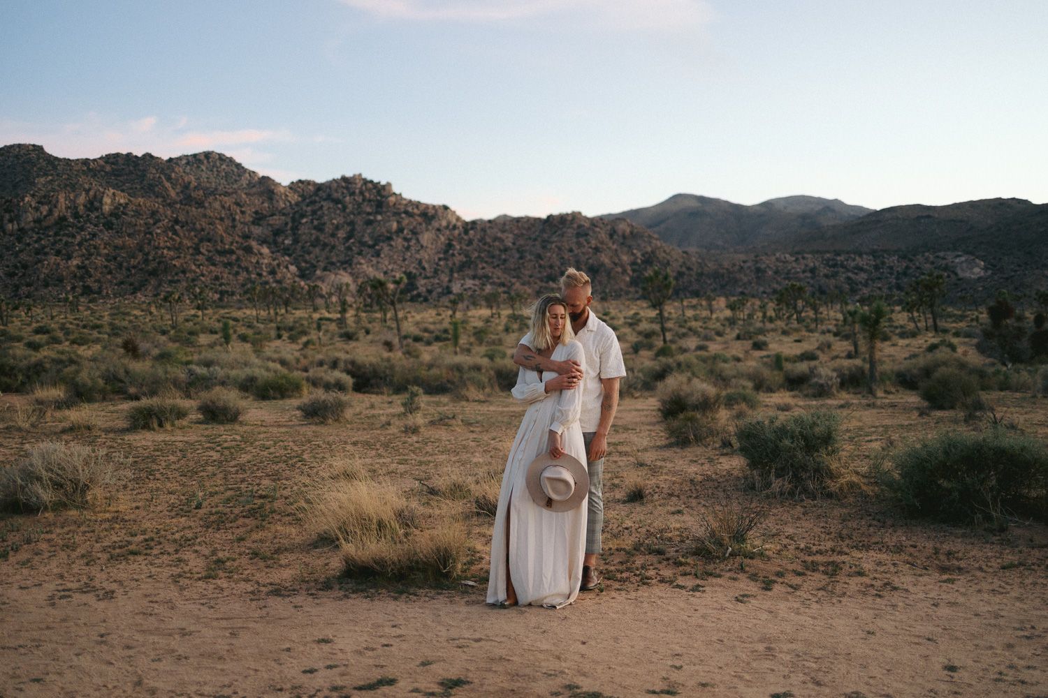 A couple in white clothing stands together in a desert setting with mountains behind them.