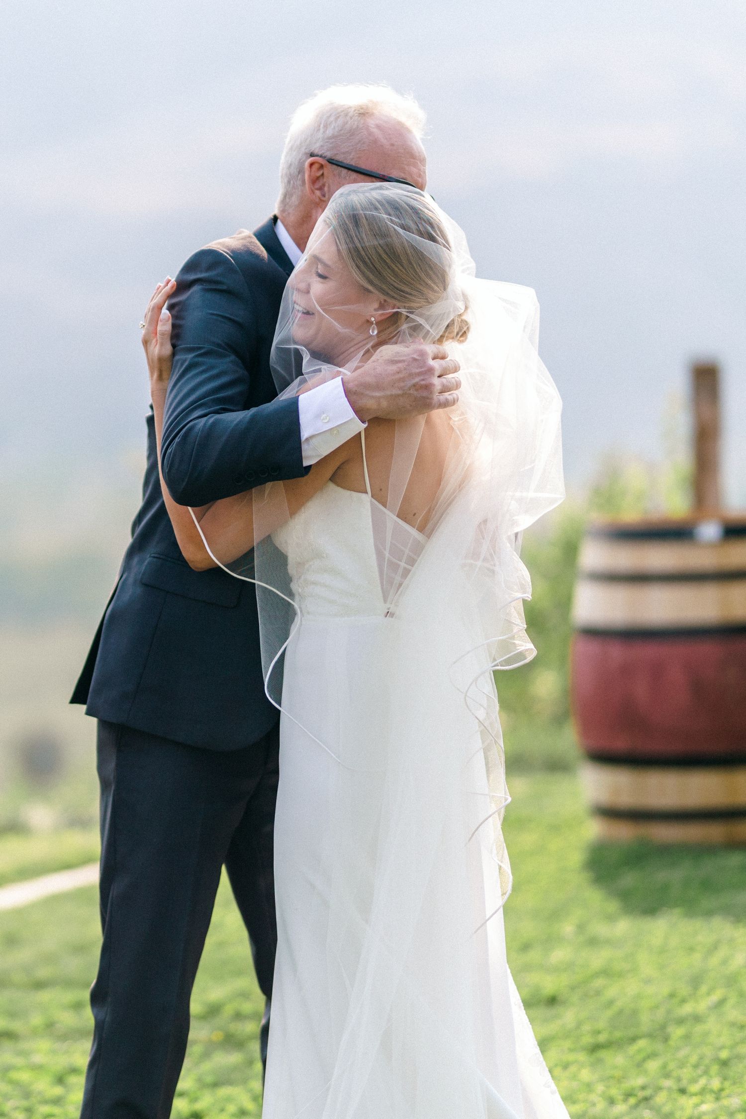 Emotional embrace between wedding couple with scenic mountain views and wine barrels in background.
