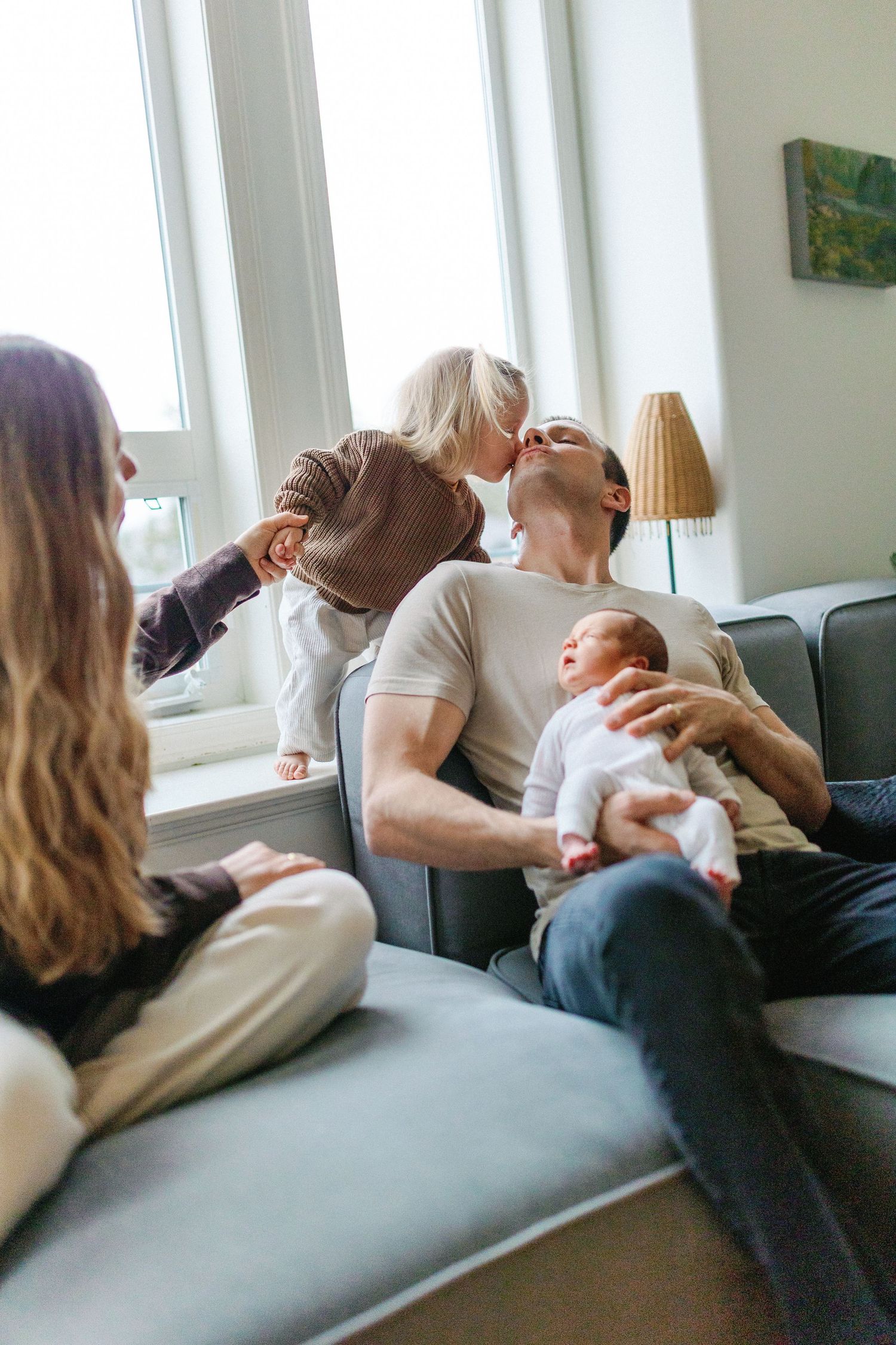 A family relaxes together on a gray couch in a bright living room with natural light streaming through windows.