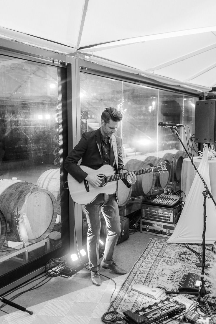 Musician performs with acoustic guitar in front of wine barrels at wedding reception venue.
