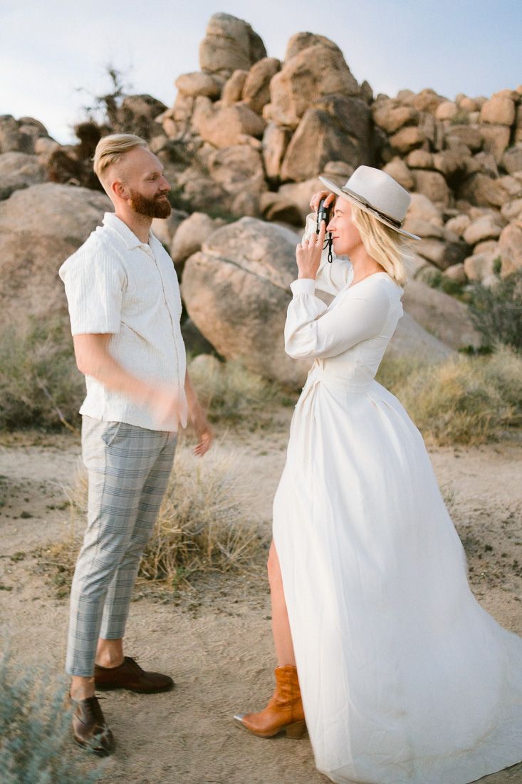 A couple poses in a desert landscape with large rock formations, wearing white and casual attire.