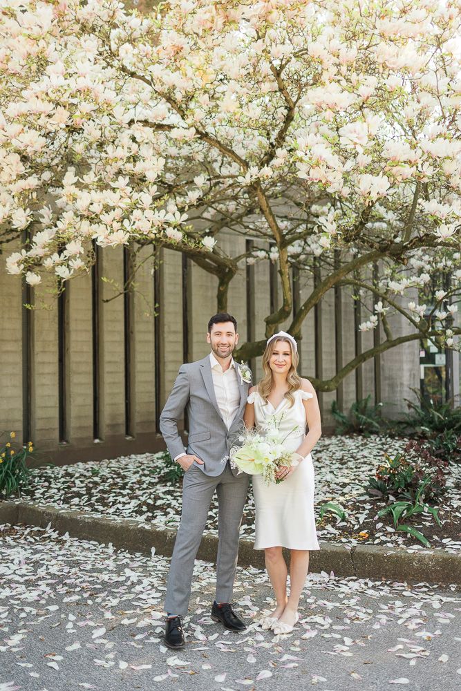 Wedding couple stands together under flowering tree with white petals scattered on ground.