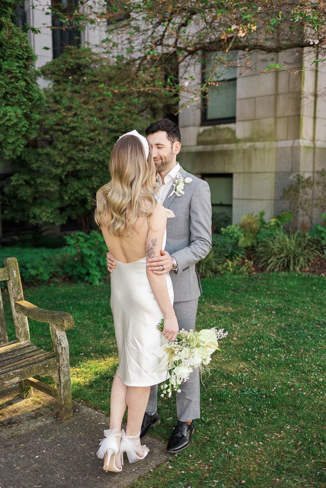 A romantic wedding moment outdoors with a couple embracing in front of greenery, the bride in white and groom in grey.