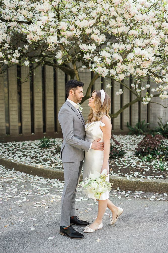 Elegant wedding couple poses beneath blooming spring trees with scattered petals.