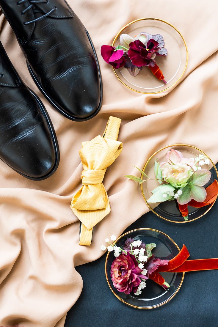 Wedding accessories including black dress shoes, yellow bow tie, and floral boutonnieres on silk fabric.