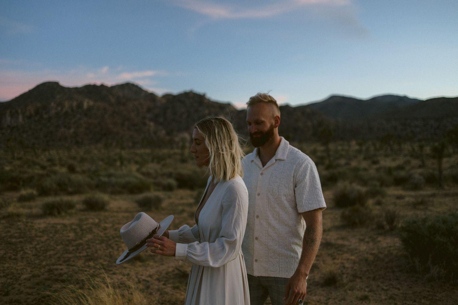 A couple stands together in a desert landscape with mountains and Joshua trees.