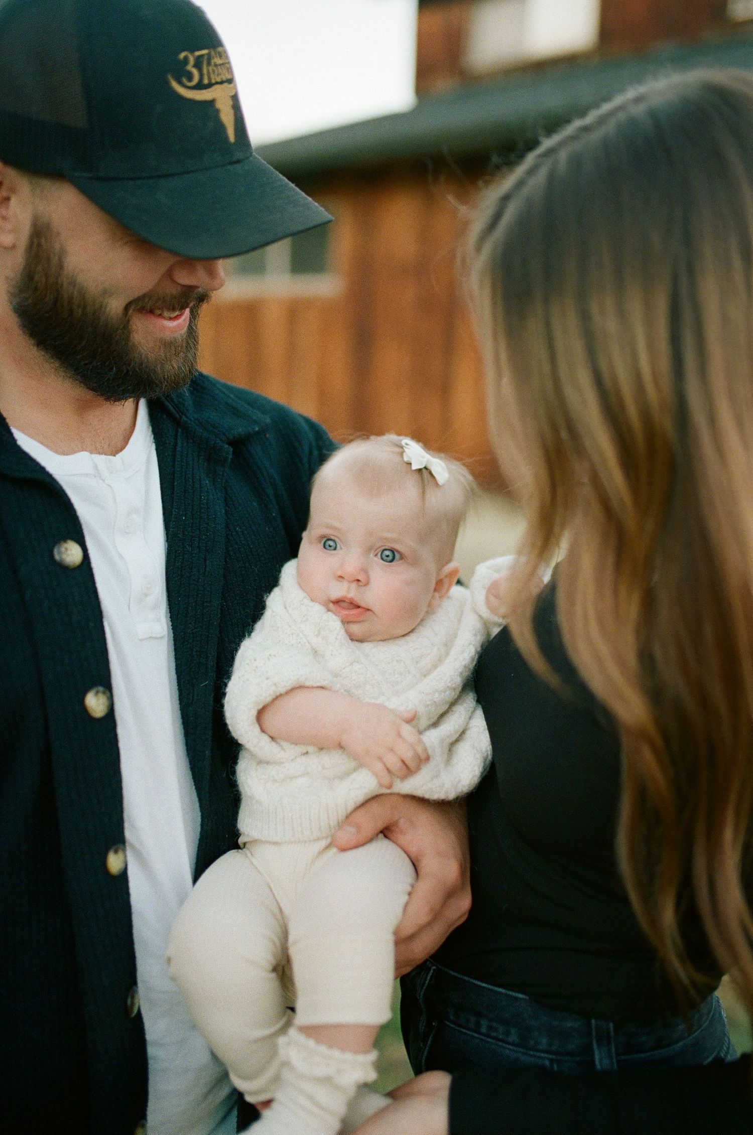A baby in a white outfit is held between two adults during a tender family moment.
