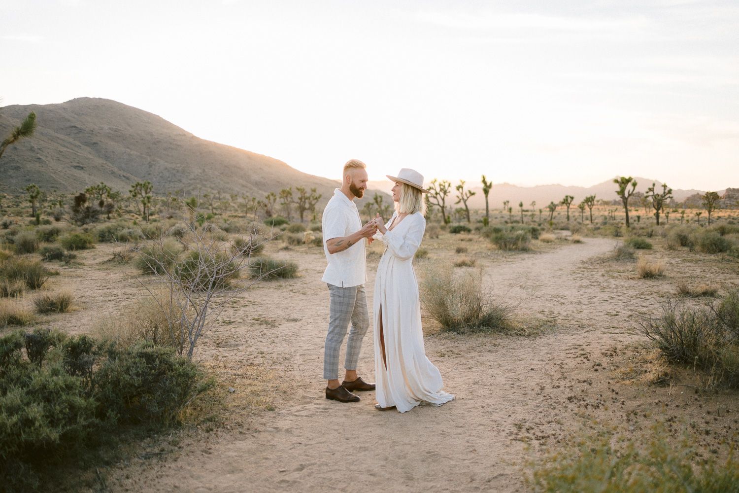 A couple embraces on a desert trail during sunset with mountains in the background.
