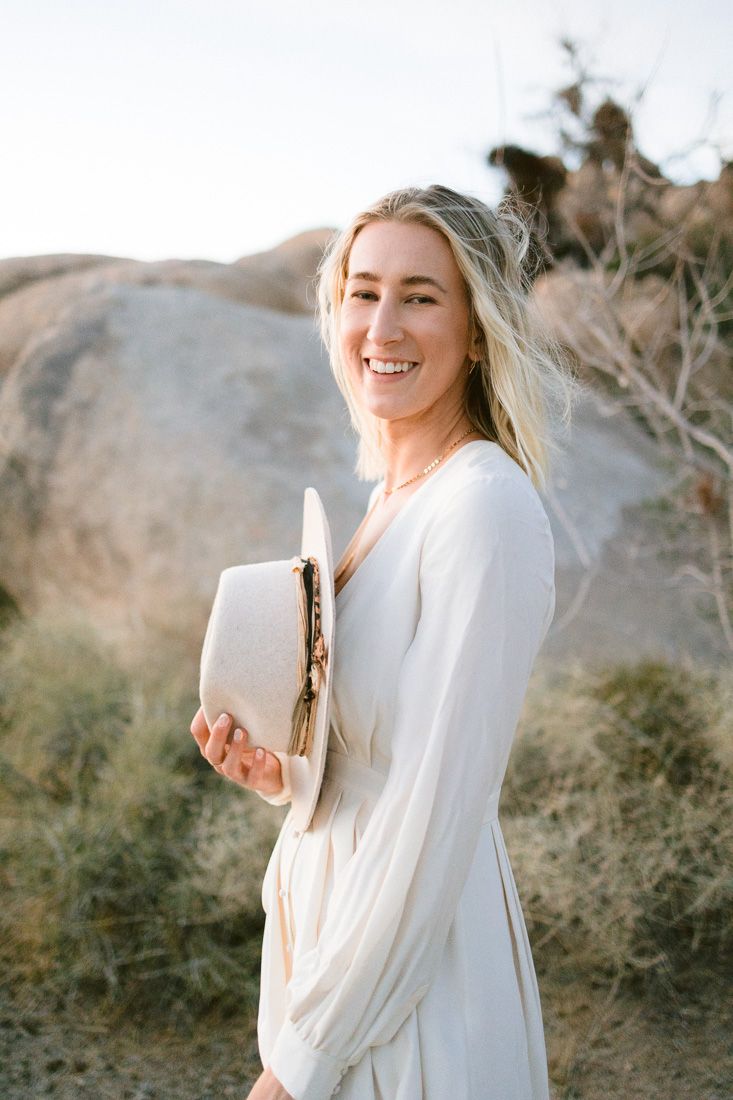 A woman in a flowing white dress smiles while holding a hat against a desert landscape with rock formations.